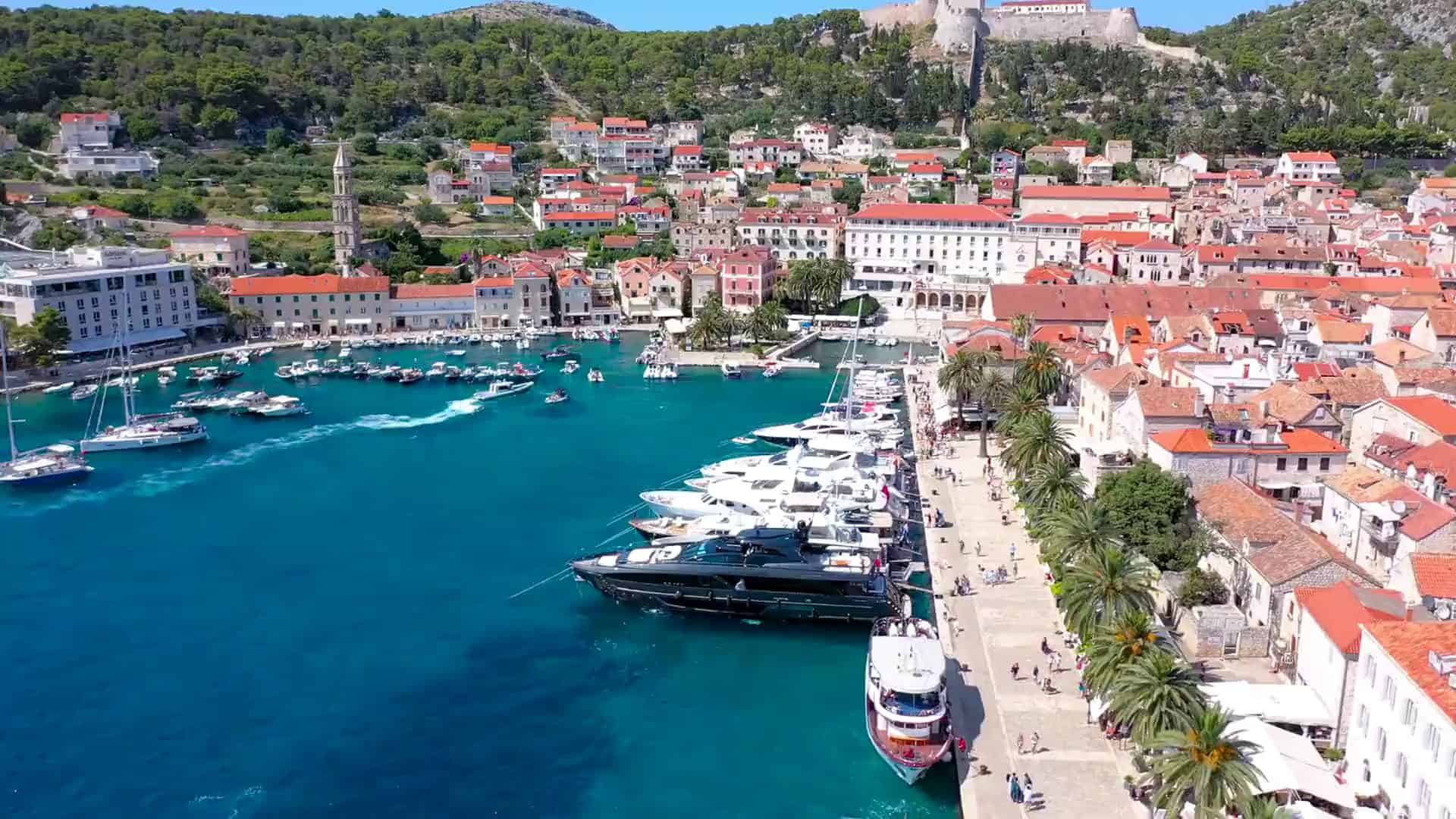 Pogled iz zraka na luku grada Hvara s jahtama uz rivu obrubljenu palmama i tvrđavom Fortica iznad starog grada