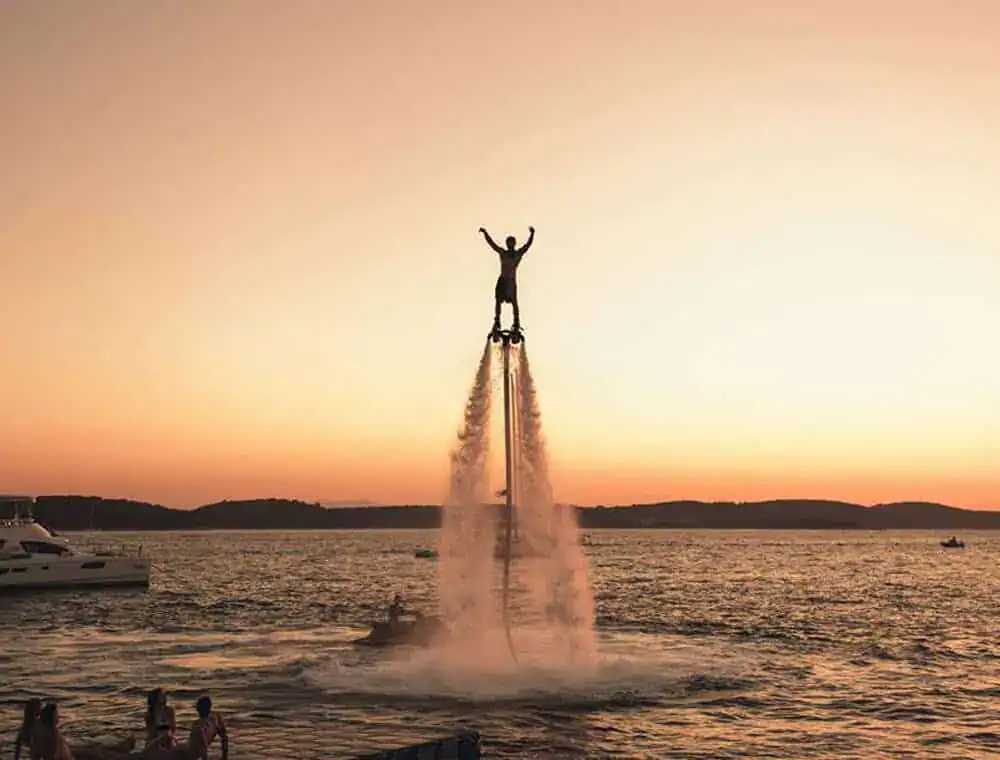 Flyboarder højt over vandet i solnedgang ud for Hvar, med en jetski og en yacht bagved
