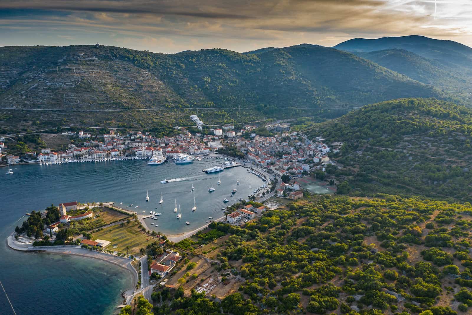 Vis town and its harbour from the air at dusk, boats anchored in the bay and the hills of Vis behind