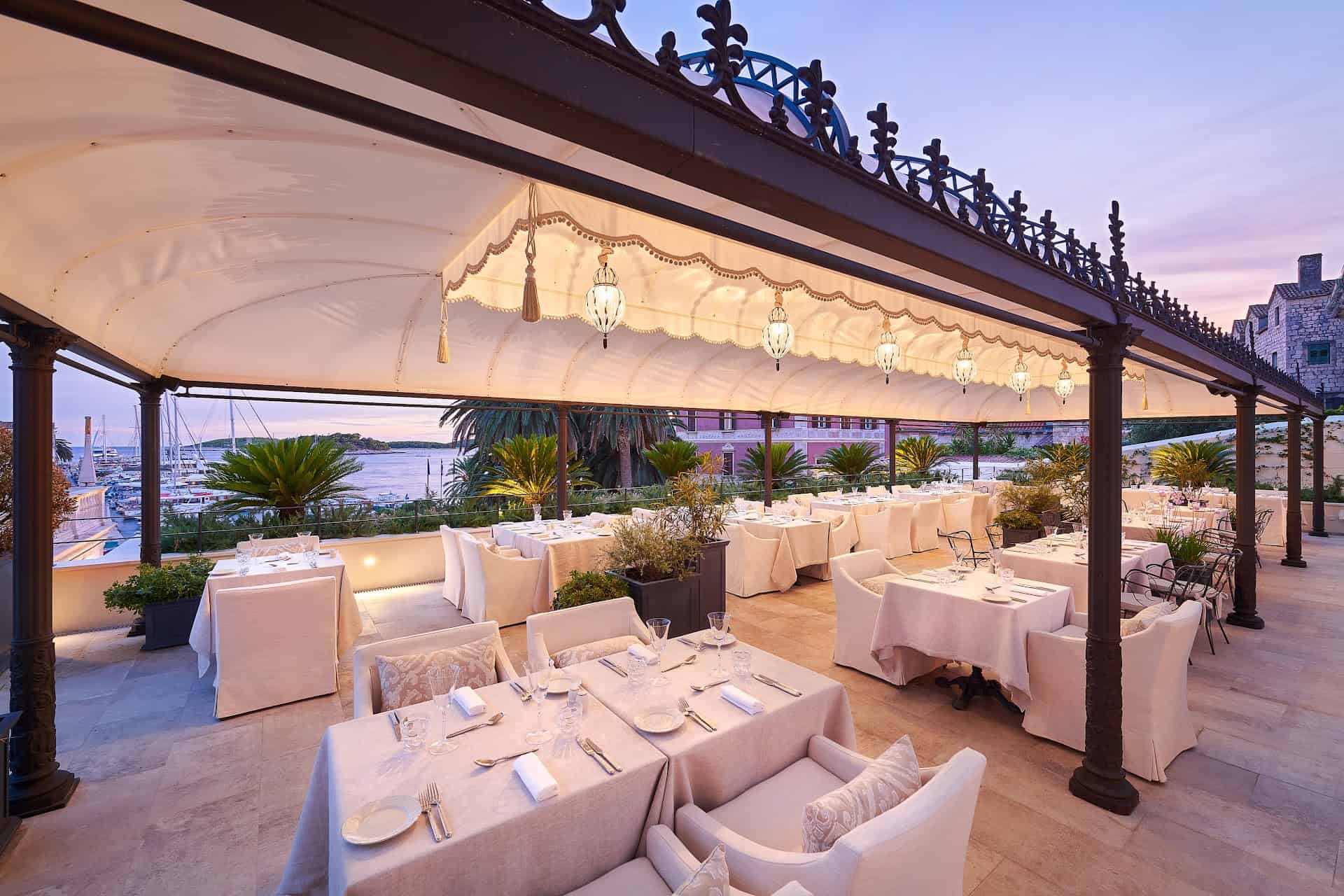 The awninged terrace restaurant of Palace Elisabeth at dusk, tables set and looking out over Hvar harbour